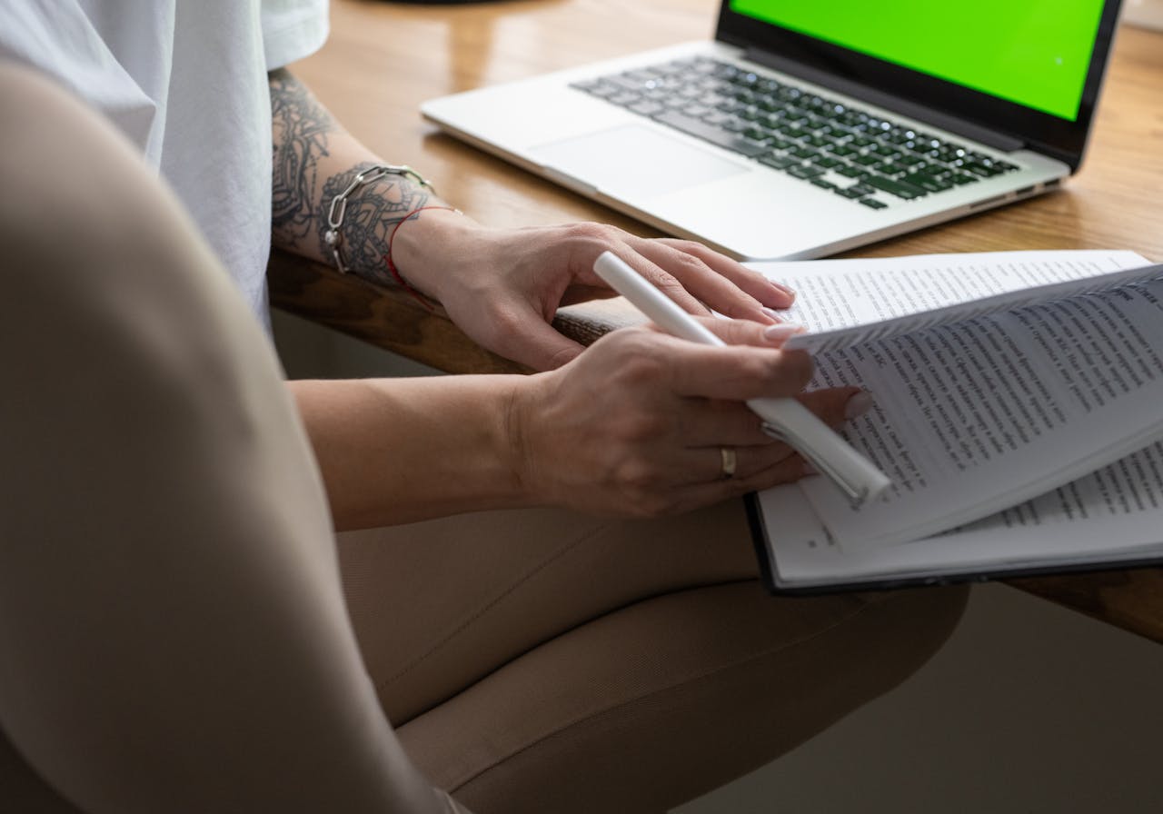 Crop faceless lady with tattoo on arm sitting on chair at table with laptop and reading book during distance lesson at home